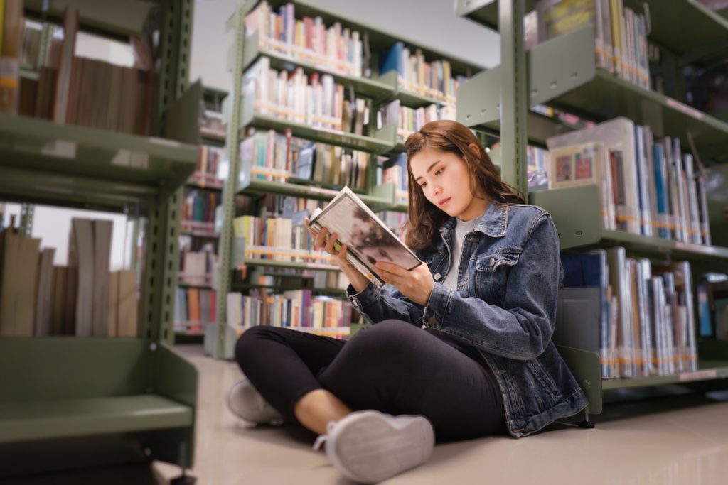 student sitting on floor in the library, Open and learning textbook from bookshelf