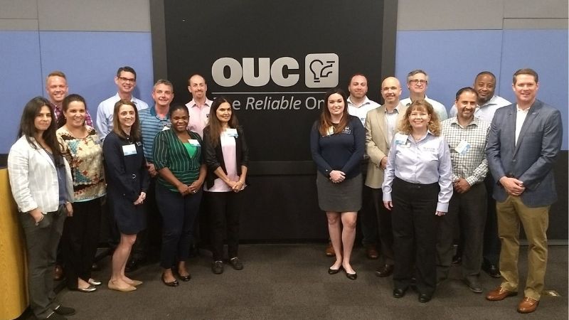 A group of students and professionals posing in front of an OUC energy company sign during network event at Crummer