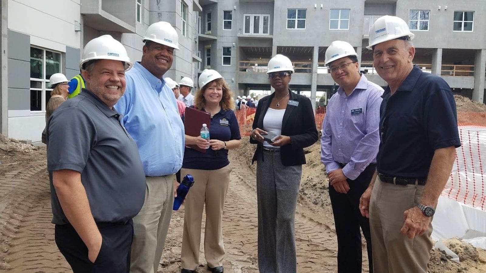 A group of smiling Crummer students and business owners wearing hard hats with an outdoor construction site in the background