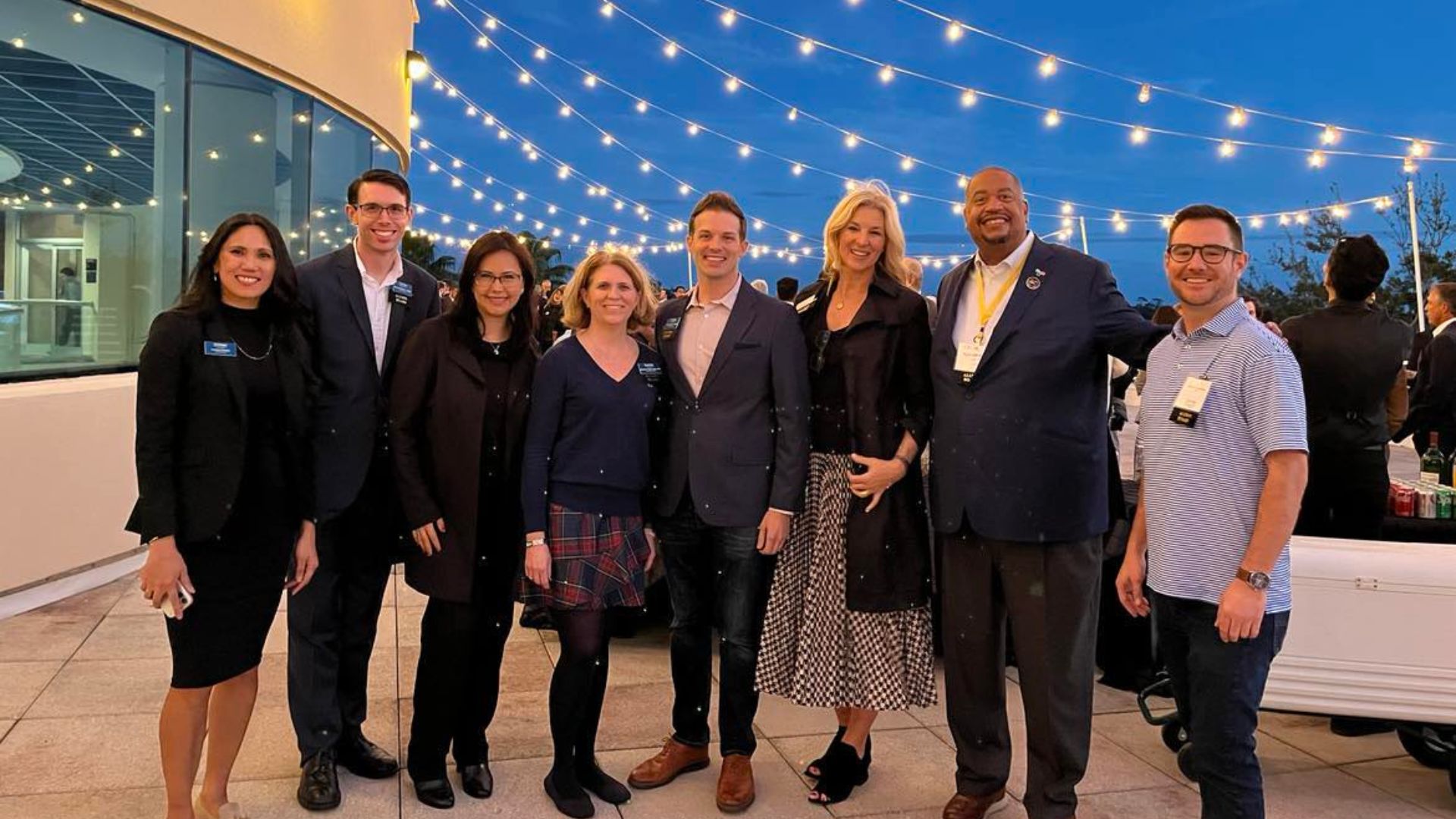 Group of eight Crummer alums and students smiling outdoors at night under string lights at Rollins College alumni event