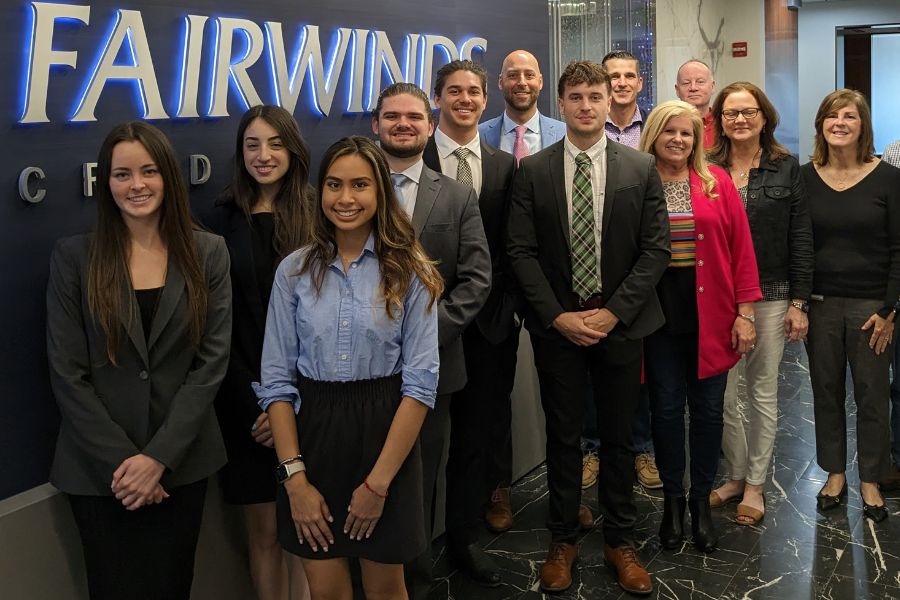 Group of Crummer Graduate School students and faculty posing in front of Fairwinds Credit Union sign, smiling at camera