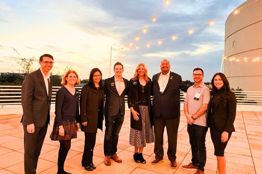 Eight Crummer Graduate School alums and students pose on rooftop terrace with string lights and sunset sky