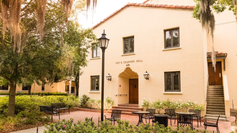 A stucco building with a sign reading Roy. E. Crummer Hall above the door is surrounded by greenery on Rollins College campus.