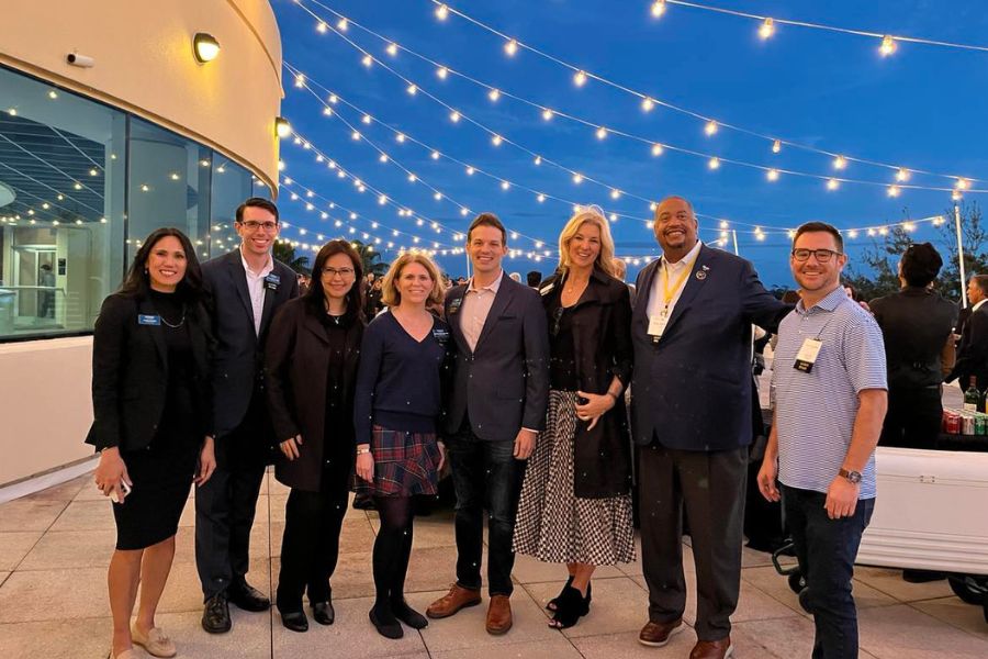 Rollins College Crummer alums stand in front of a dark evening blue sky at an alumni event, string lights visible above them