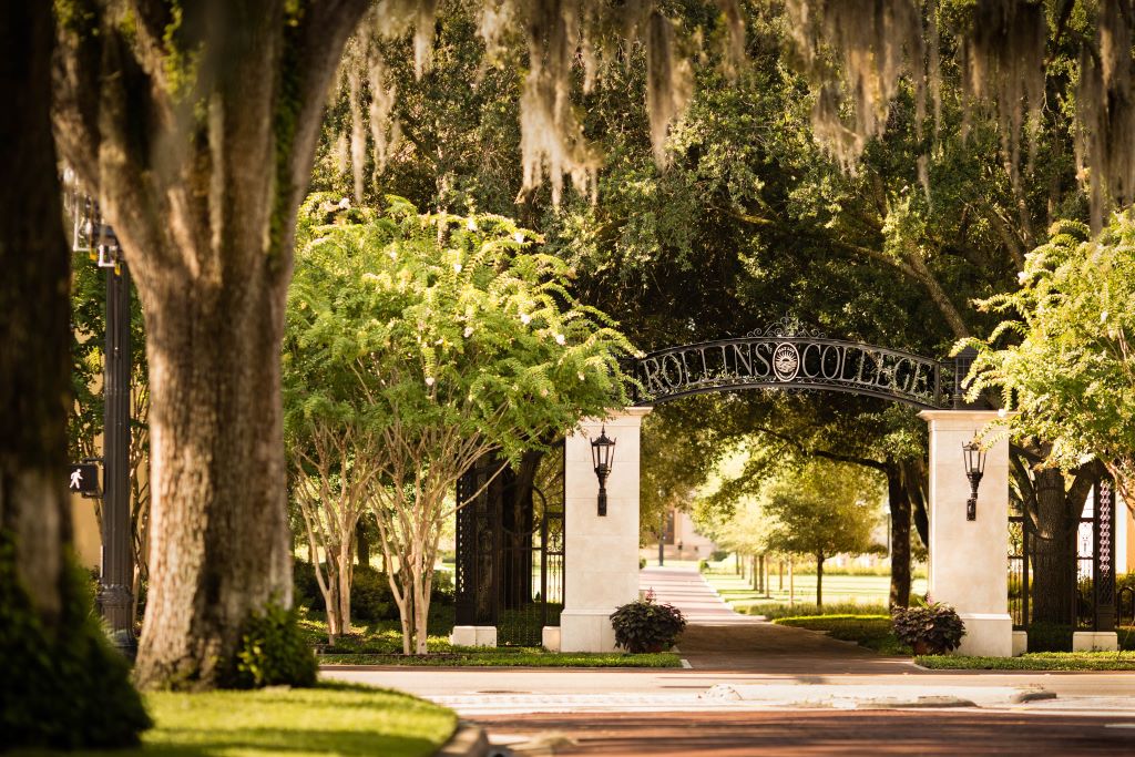 Arch reads "Rollins College" above a brick path at the Graduate School of Business campus entrance surrounded by lush trees