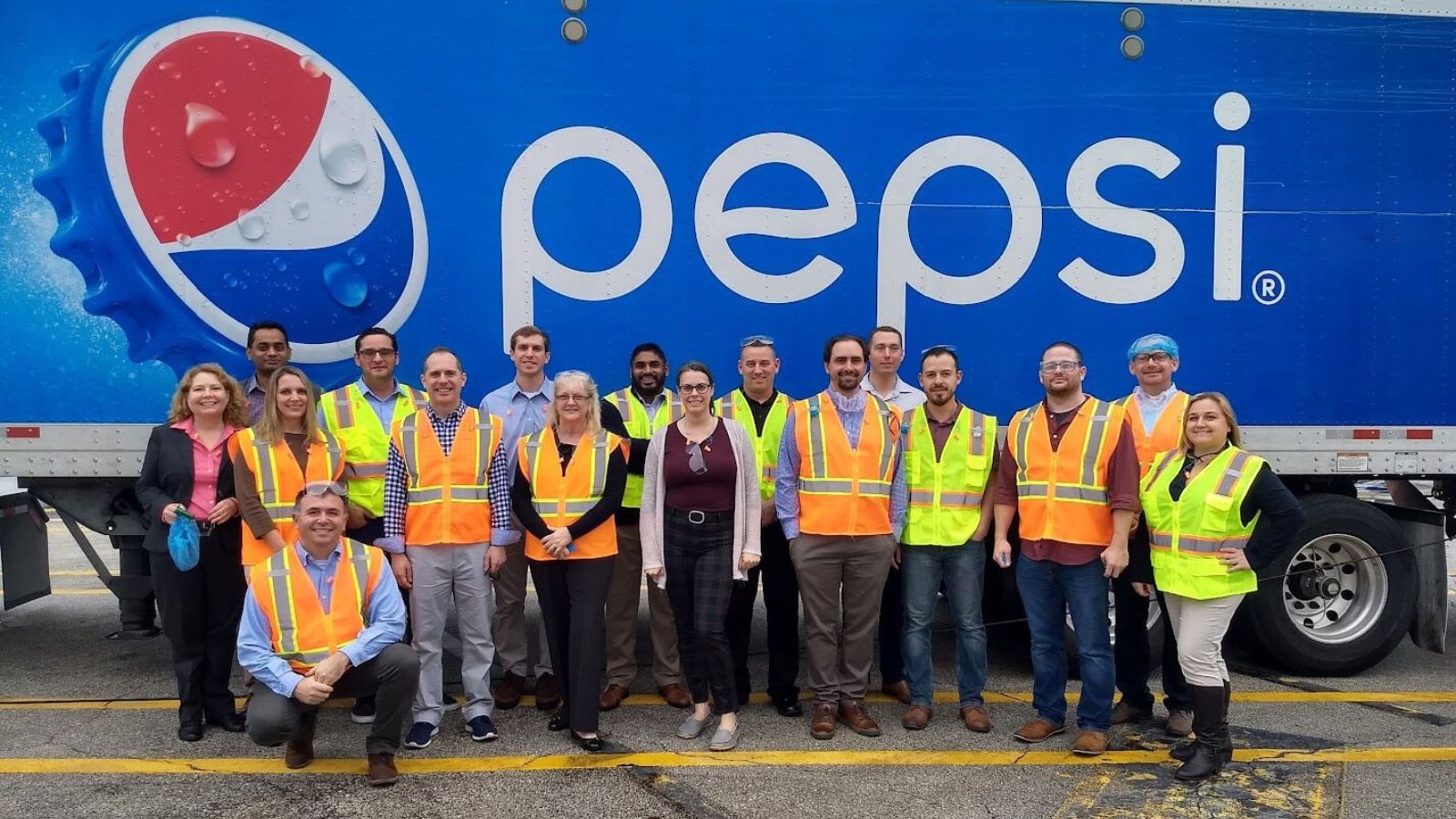 Crummer MBA students in orange and yellow safety vests pose with Pepsi employees in front of a blue Pepsico 18-wheeler truck