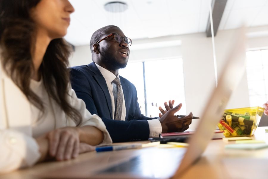 Graduate business students at table with laptop listen to gesturing professor, woman adds notes to glass board in background