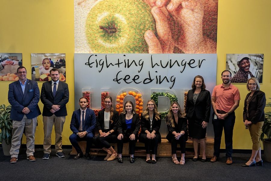 Group of students and professionals from Crummer pose in front of wall reading "Fighting hunger feeding hope" at a food bank