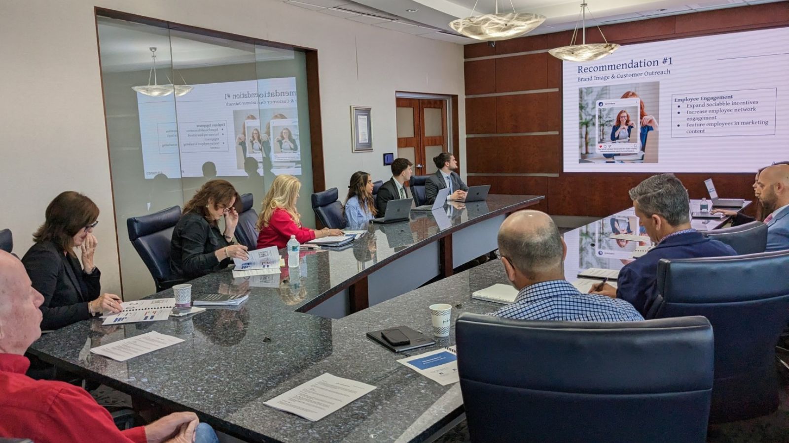 Professionals sitting at a boardroom table looking at presentation slides visible on a screen outlining brand recommendations