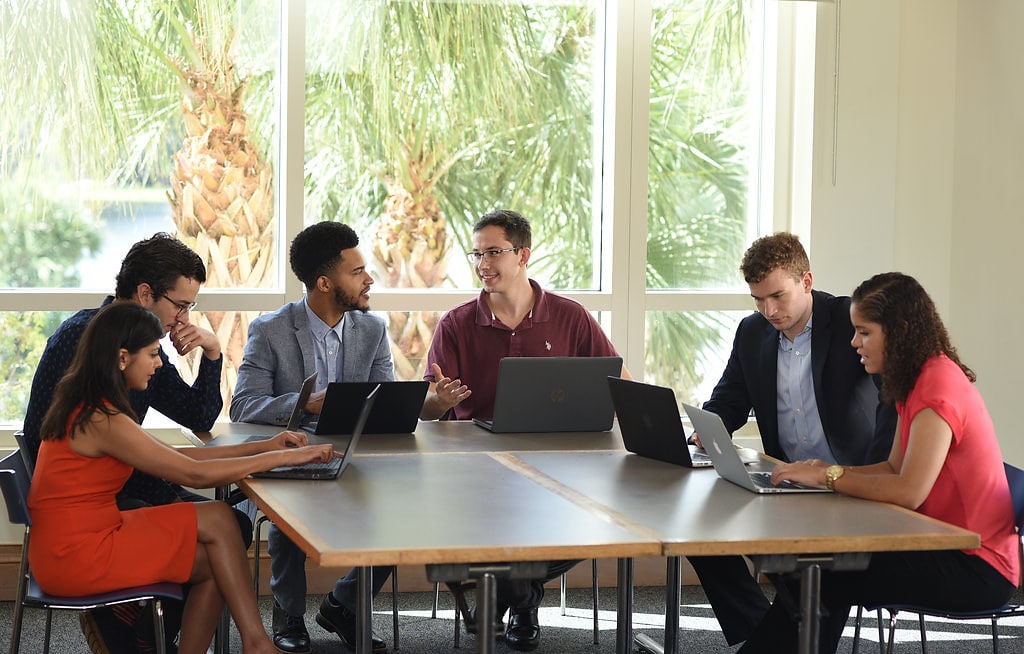 Early career MBA students at Rollins Crummer sit around table with laptops, palm trees visible through windows behind them