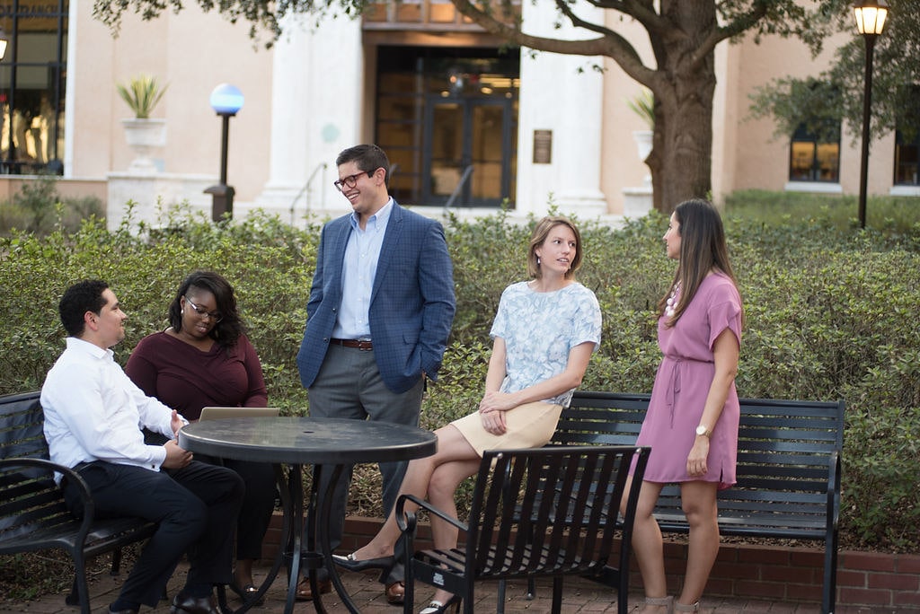 Five professional students sit and discuss at outdoor table, Bush Science Center and trees framing the Crummer campus patio