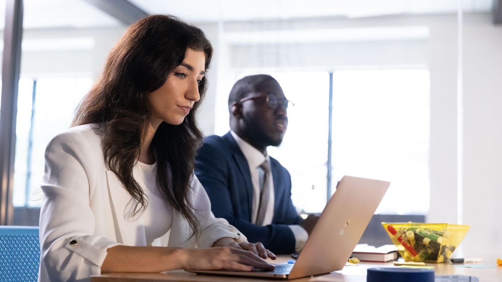 Two professionally dressed people work on a laptop and speak to someone out of frame in a modern conference table setting