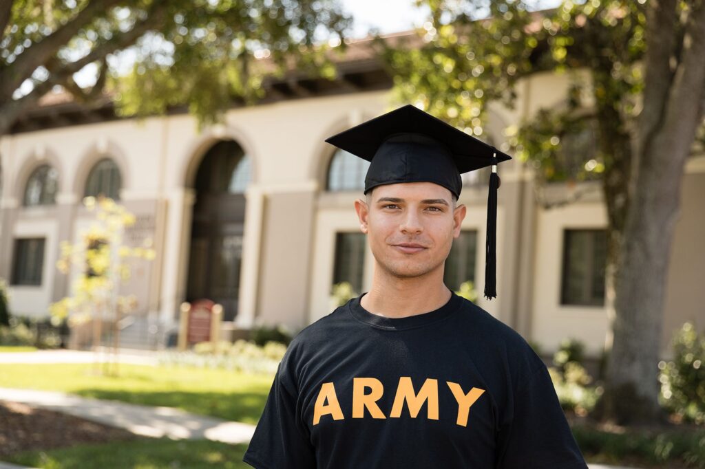 Smiling graduate in cap and Army t shirt standing in front of Spanish-style campus building with trees looking at the camera