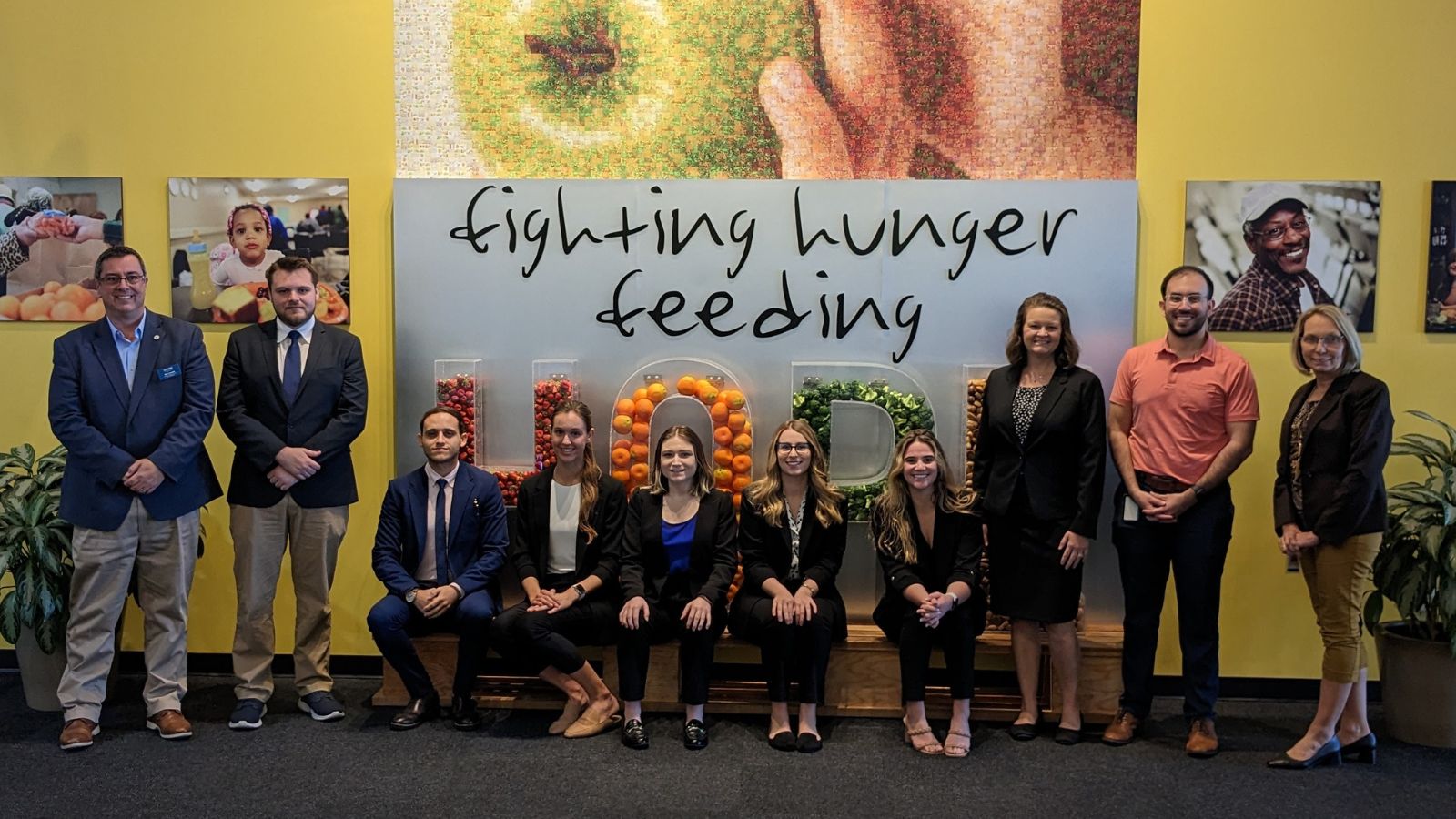 Group of students and business professionals stand in front of a display wall reading "fighting hunger, feeding hope"