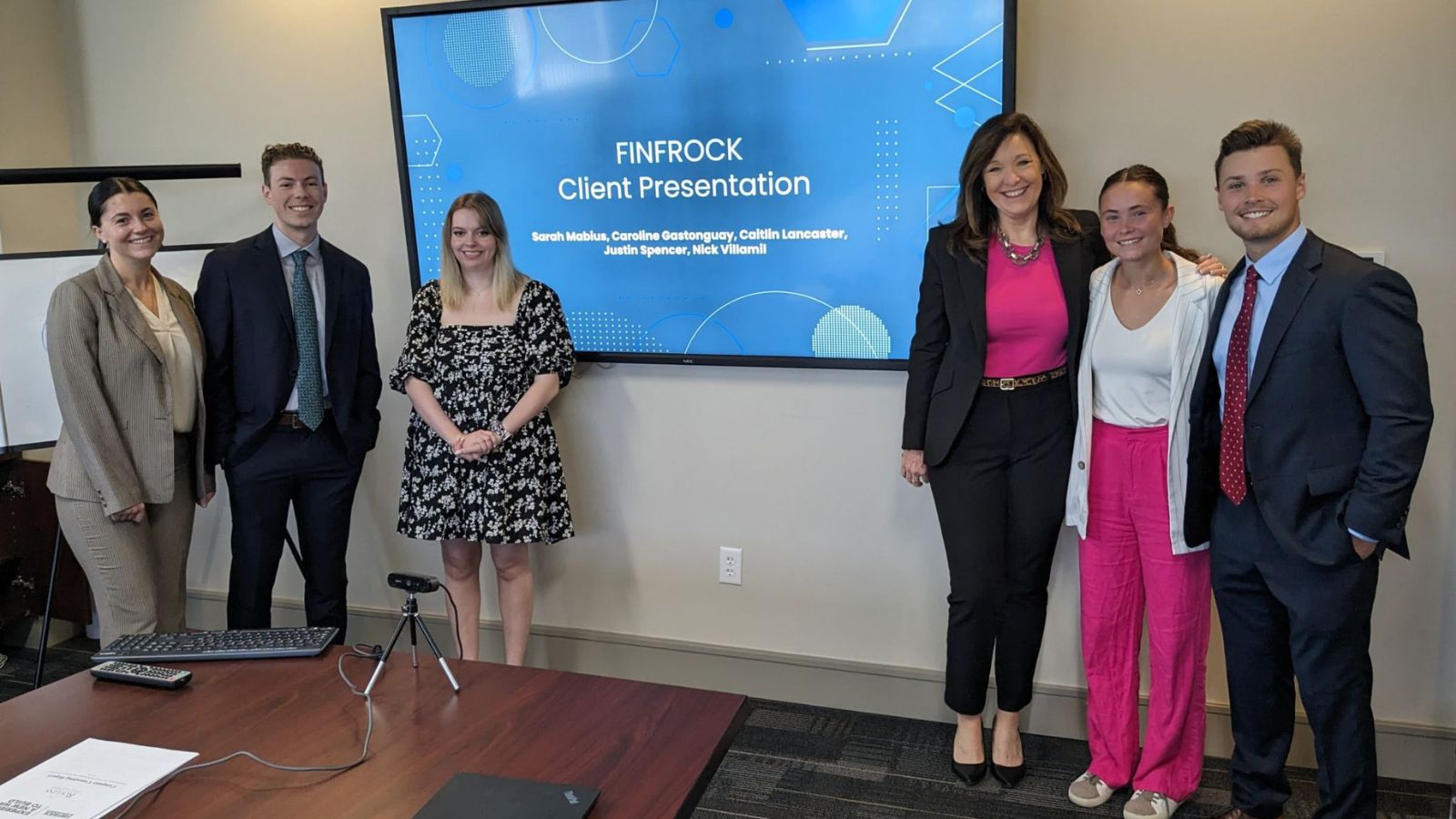 Six Crummer students stand near a conference table in front of a screen showing a slide reading "Finrock Client Presentation"