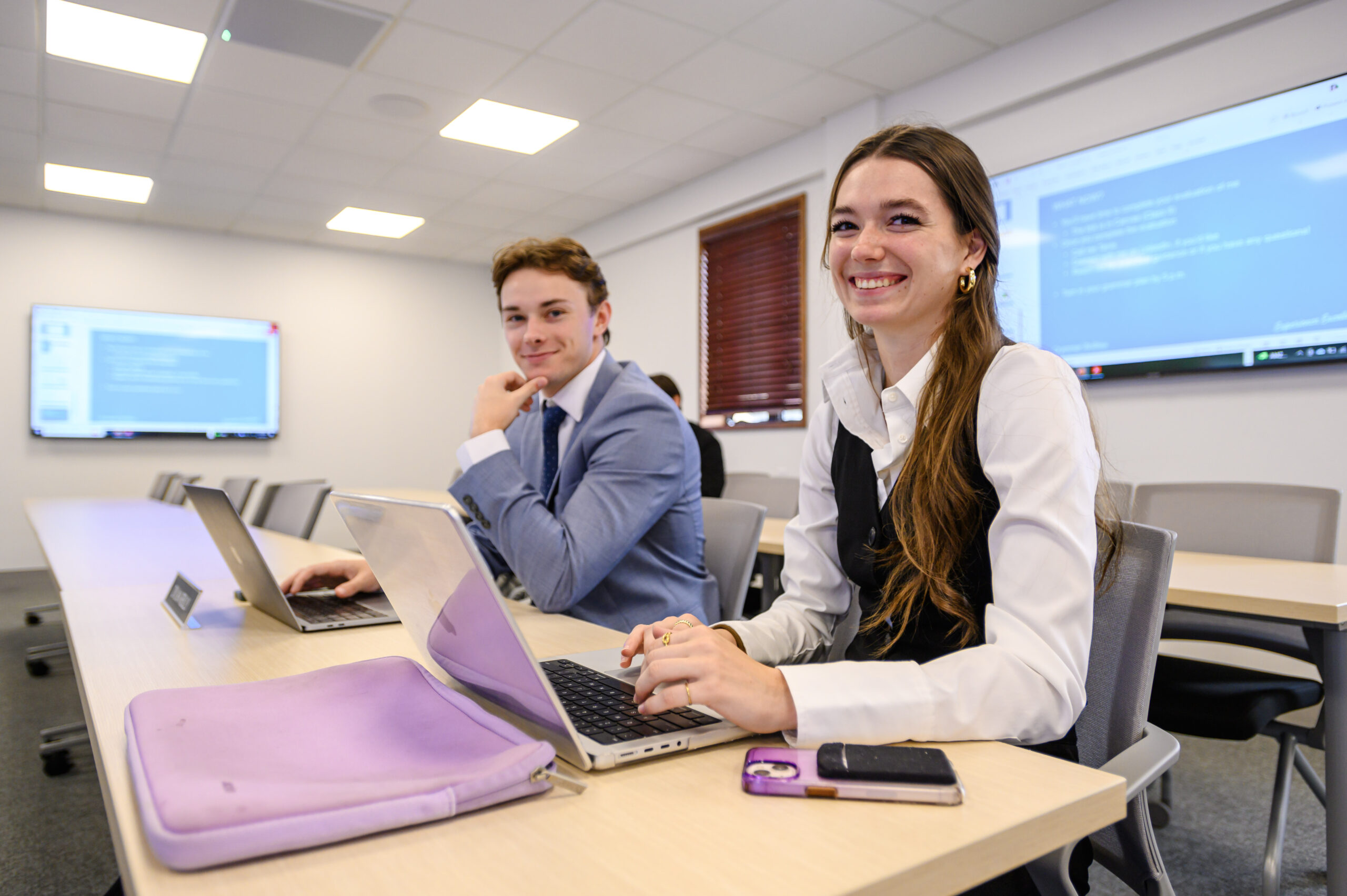 one female student smiles and types on her computer next to a male student who has his hand on his chin