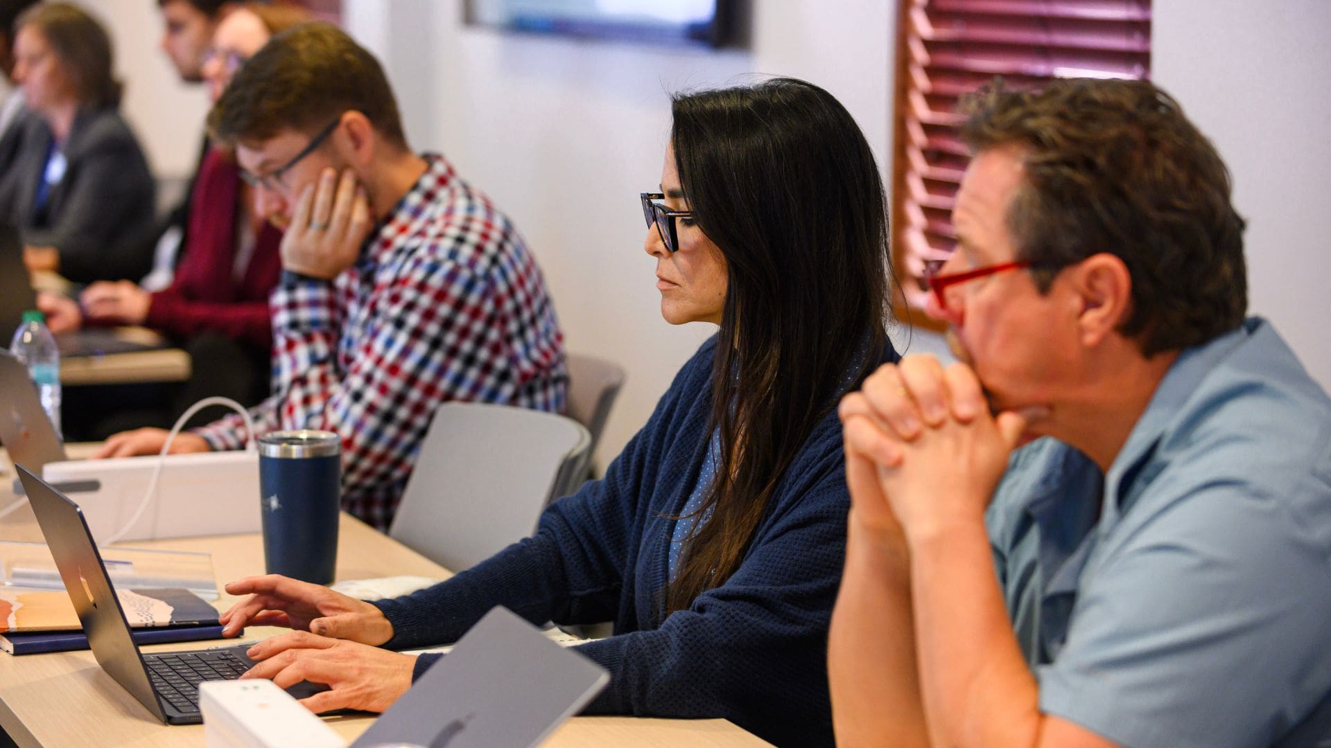 adult female with long straight brown hair and glasses sitting in a classroom typing on her laptop