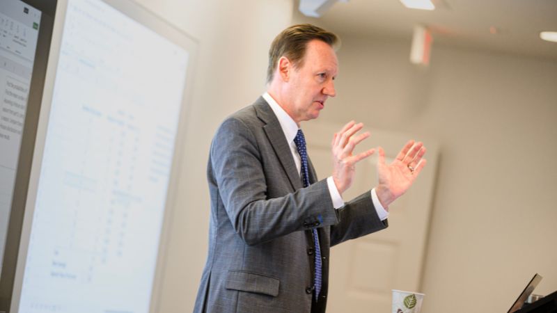 professor keenan yoho in a gray jacket and blue tie in the front of the class motioning with his hands