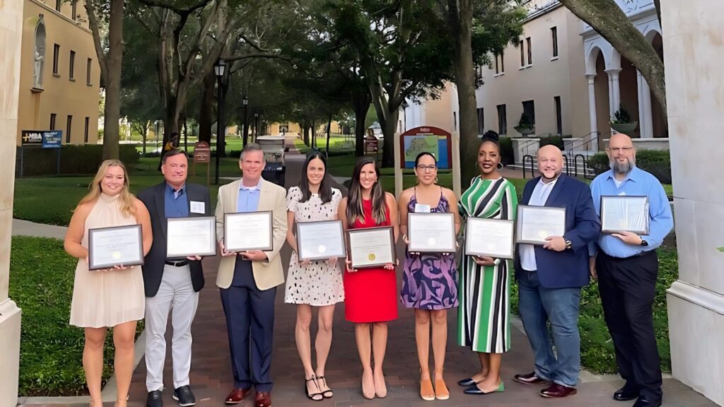 a group of adults hold crummer certificates while standing on a brick pathway between buildings