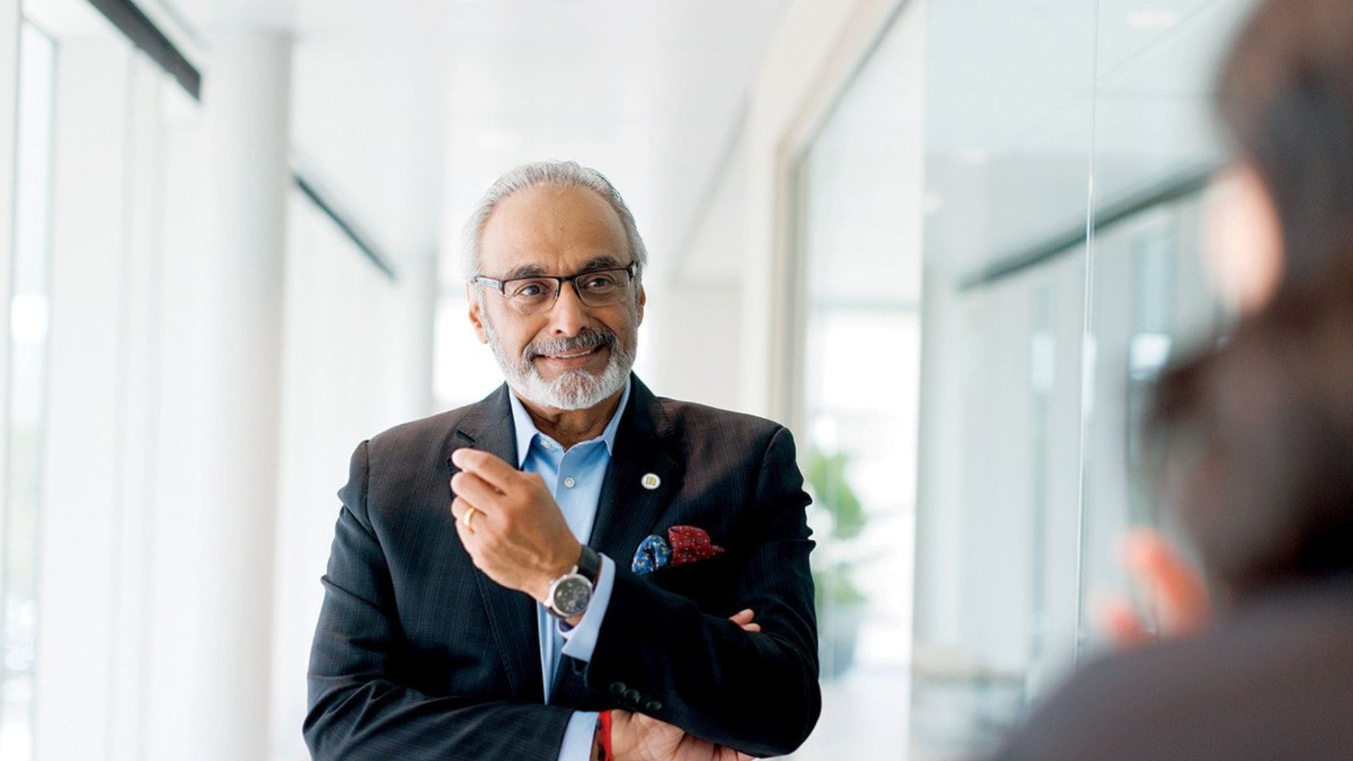 dean anil menon in a room with lots of light holding up his arm as he smiles at another person not pictured