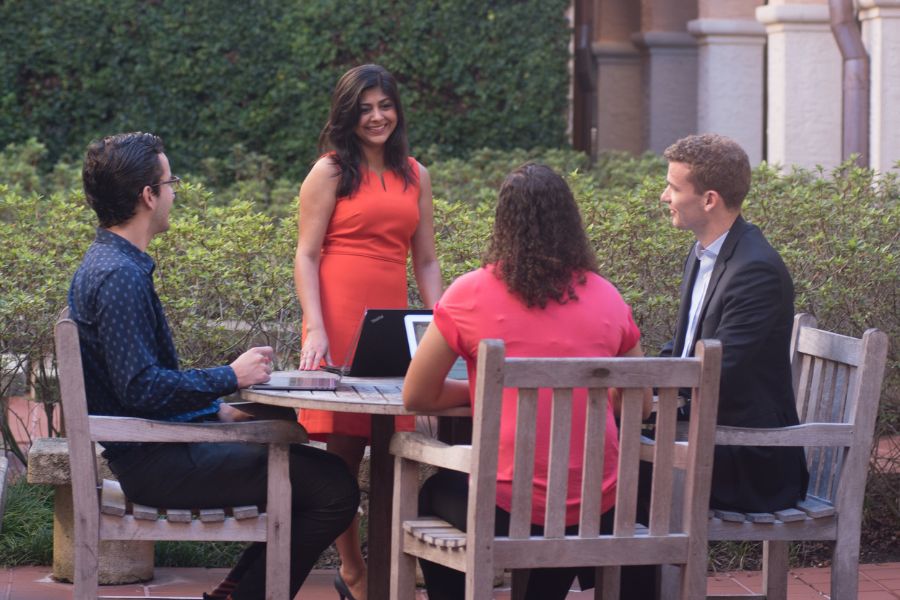 four students sit outside at a table with one female student standing leading the group