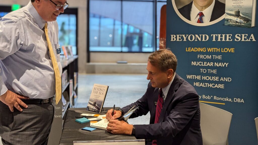 bob roncska sits at a table and signs his book for crummer staff mike kazazis