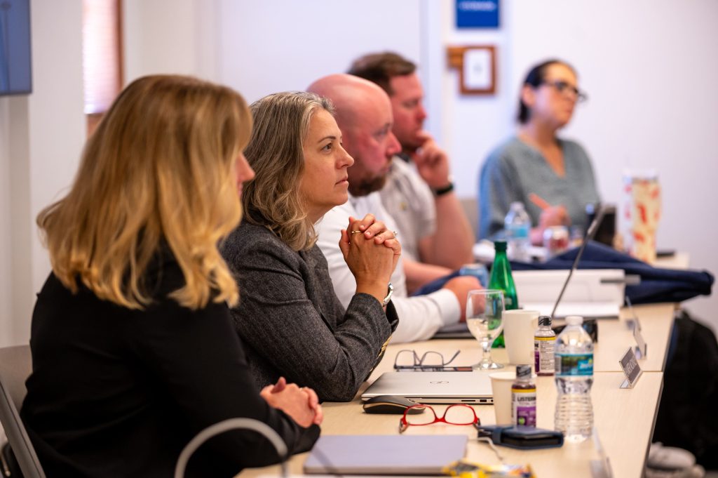 A panel of three people, including a woman in the foreground, seated at a long table during a focused professional or academic meeting.