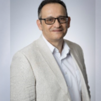 Professional headshot of a man wearing black glasses, a light beige blazer, and a white collared shirt against a light gray background.